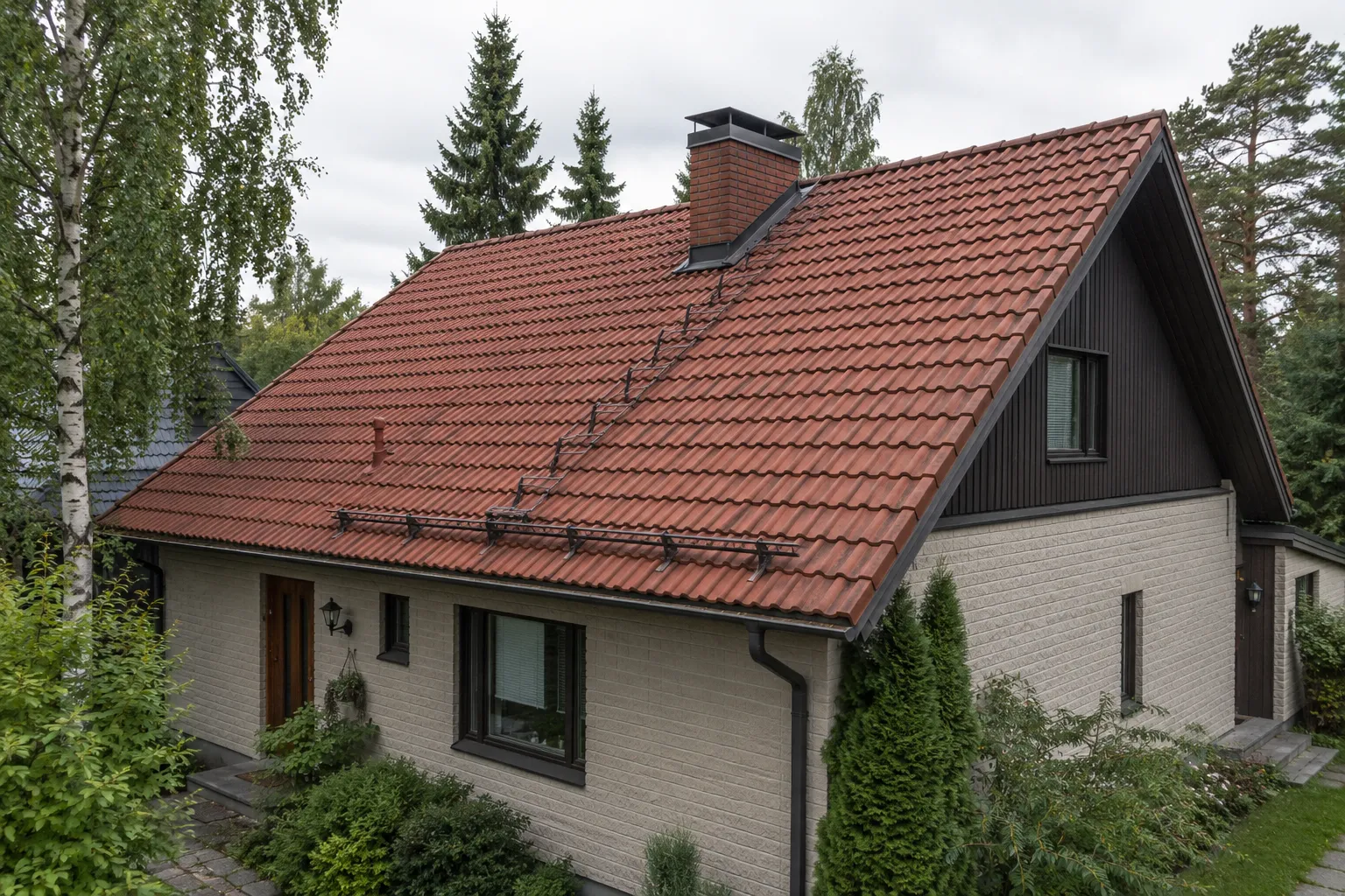 Vaaleatiilinen omakotitalo, jonka jyrkkä harjakatto on tiilenpunaista betonitiiltä, katolla muurattu savupiippu ja kattosilta.