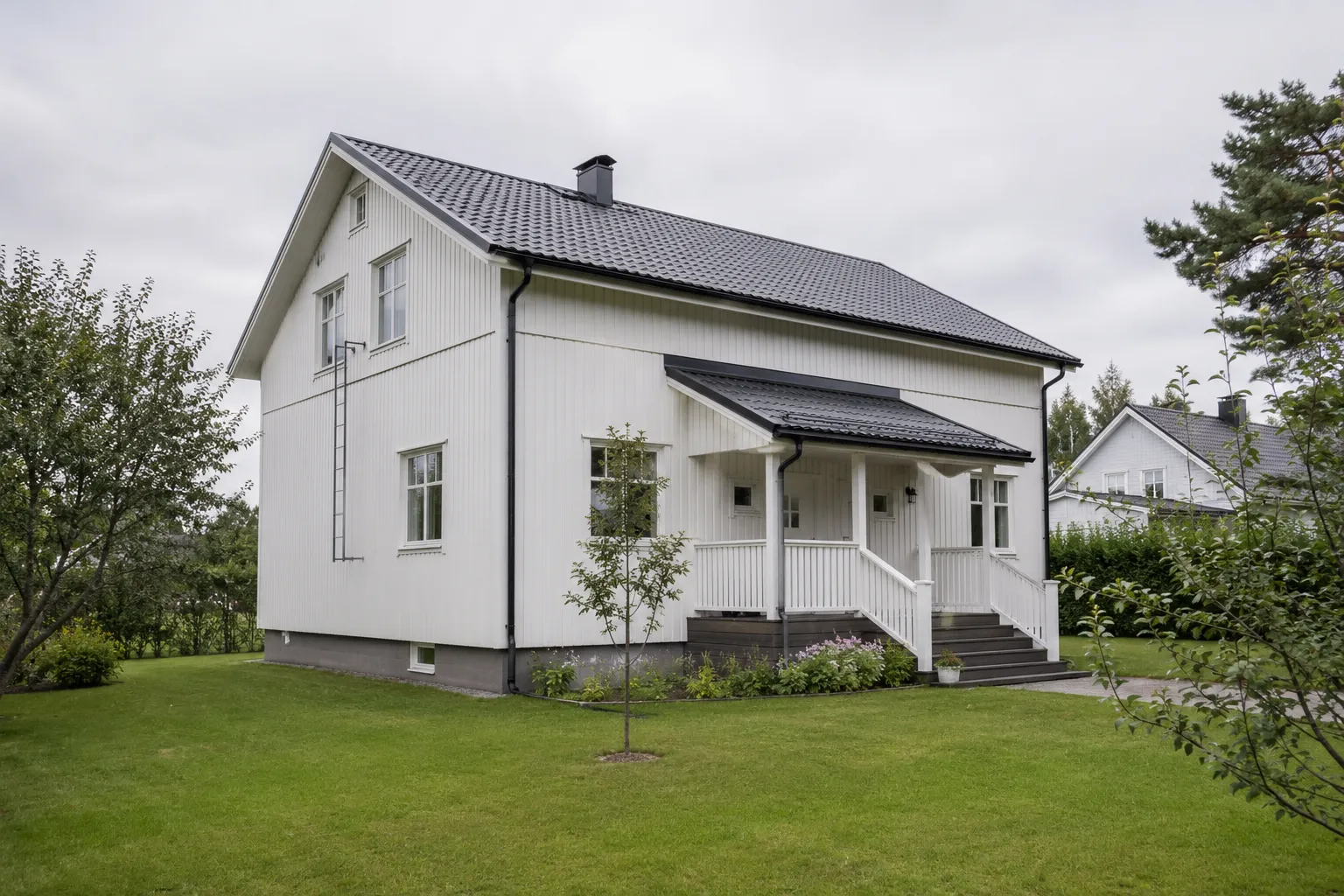 Valkoiseksi maalattu kaksikerroksinen puutalo, pystylaudoitus ja tumma katto, vihreä piha.